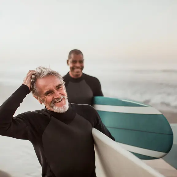 Två glada surfare i våtdräkter på stranden med surfbrädor i handen, solnedgång i bakgrunden