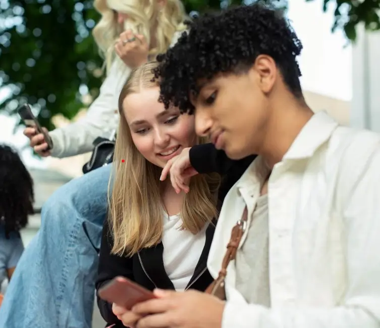 Ungdomar som använder smartphones utomhus, fokus på två personer som delar skärm och skrattar. Socialt umgänge och teknik