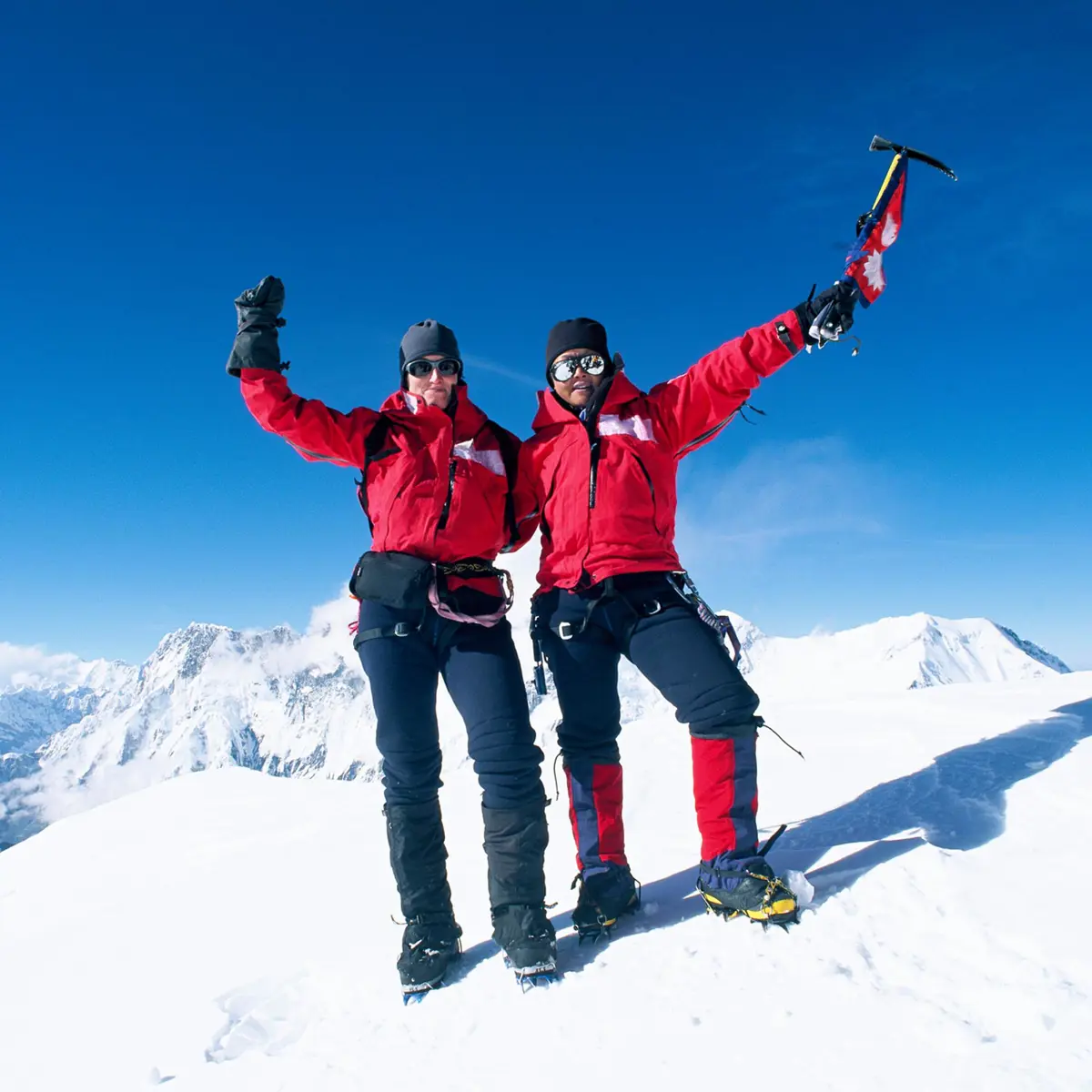 Två glada bergsklättrare i röda jackor firar på snötäckt bergstopp under klarblå himmel