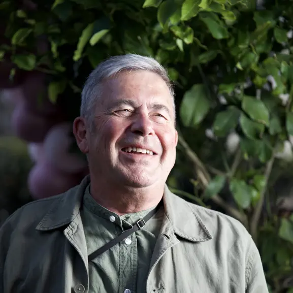 Man som står utomhus, ler och bär en grön jacka med gröna löv i bakgrunden