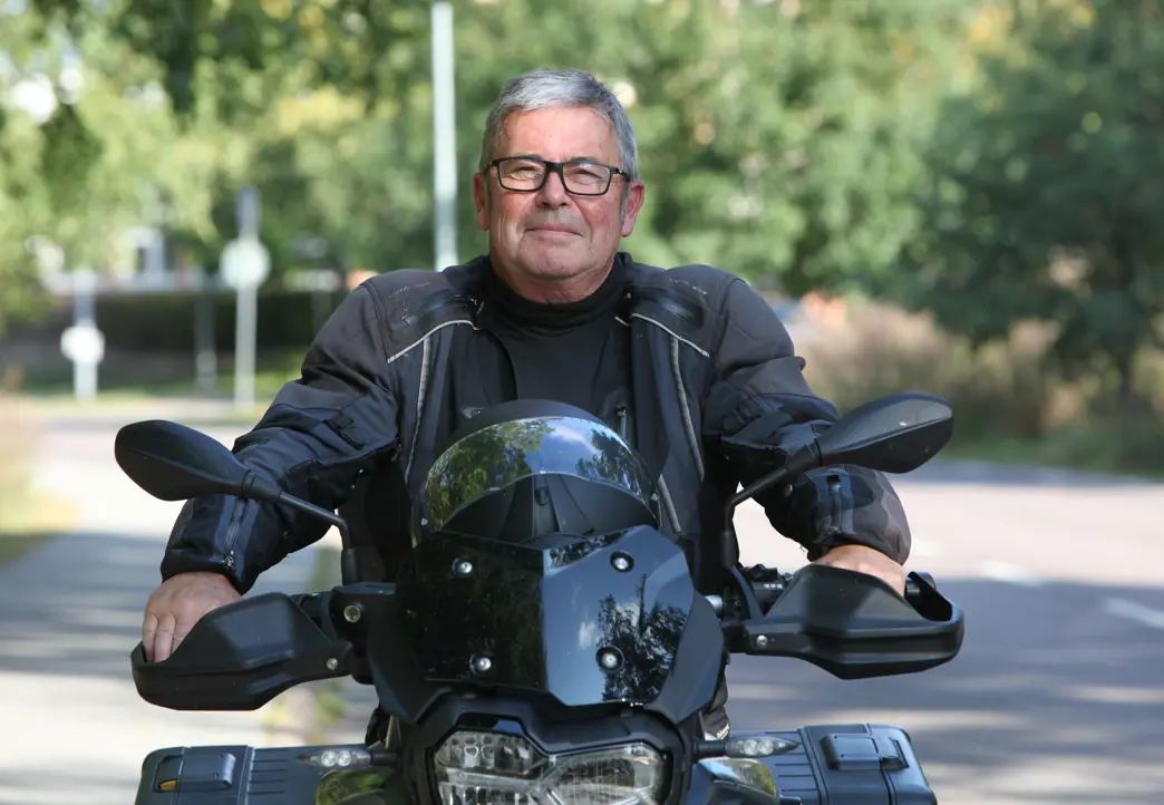 Äldre man kör motorcykel på solig landsväg, iklädd motorcykeljacka och glasögon, med grönskande bakgrund