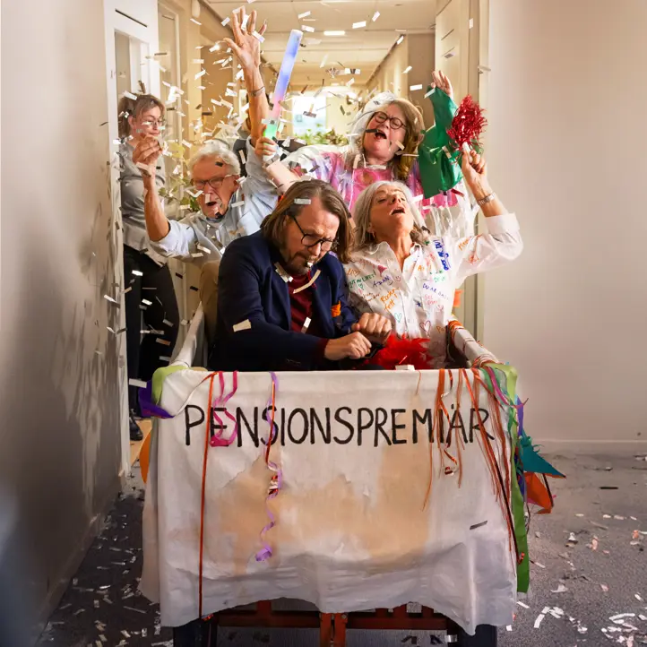 Flera personer firar pension i en dekorerad vagn, omgiven av konfetti och feststämning i en korridor
